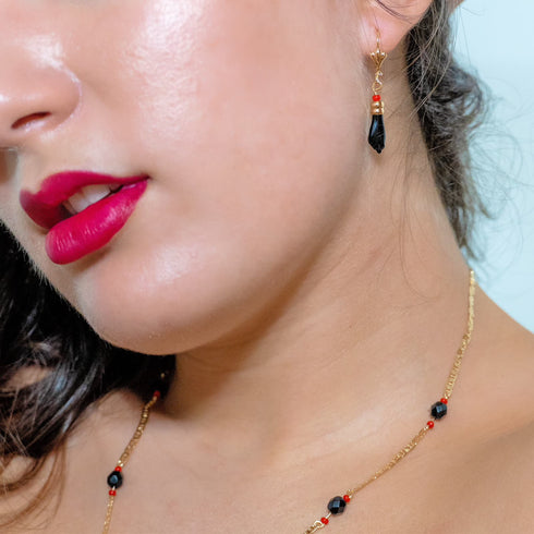 Close-up of an individual wearing a gold and black beaded Unisex Azabache Necklace Evil Eye Protection, complemented by matching earrings, with vibrant red lipstick and shiny skin.